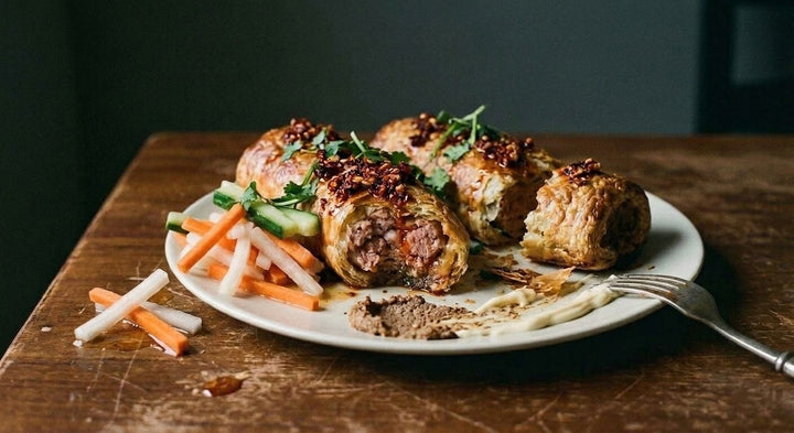 Two long banh mi pork sausage rolls on a plate, one bitten open, chilli crunch spooned over, pickled daikon and carrot, cucumber and pate to the side